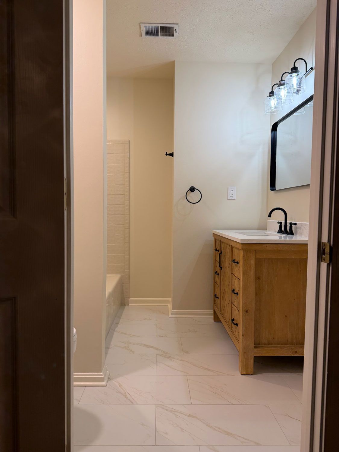 Modern bathroom with white tile floor, wooden vanity, mirror, and tub visible through doorway Modern bathroom with white tile floor, wooden vanity, mirror, and tub visible through doorway