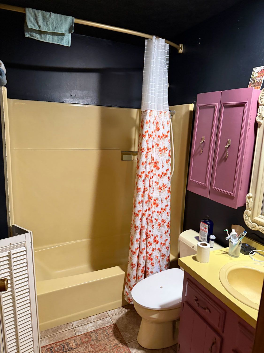 Small bathroom with tan tub, floral shower curtain, pink cabinets, white toilet, and yellow sink vanity.