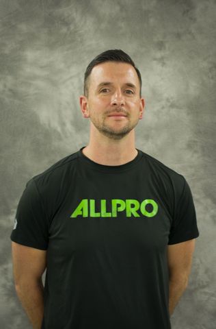 Man standing against a gray backdrop, wearing a black ALLPRO T-shirt with green lettering.