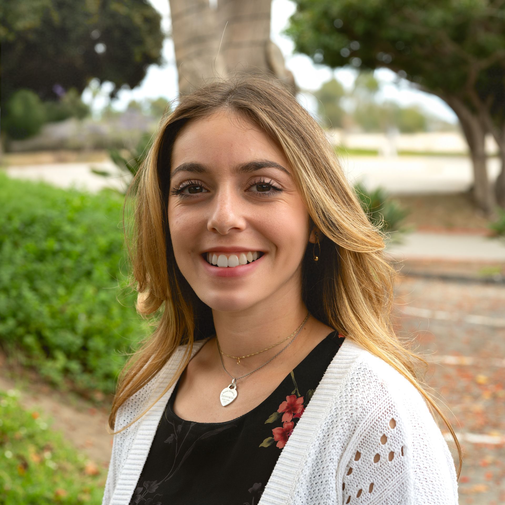 Woman smiling, wearing a white cardigan and black dress, necklace, outside near trees.