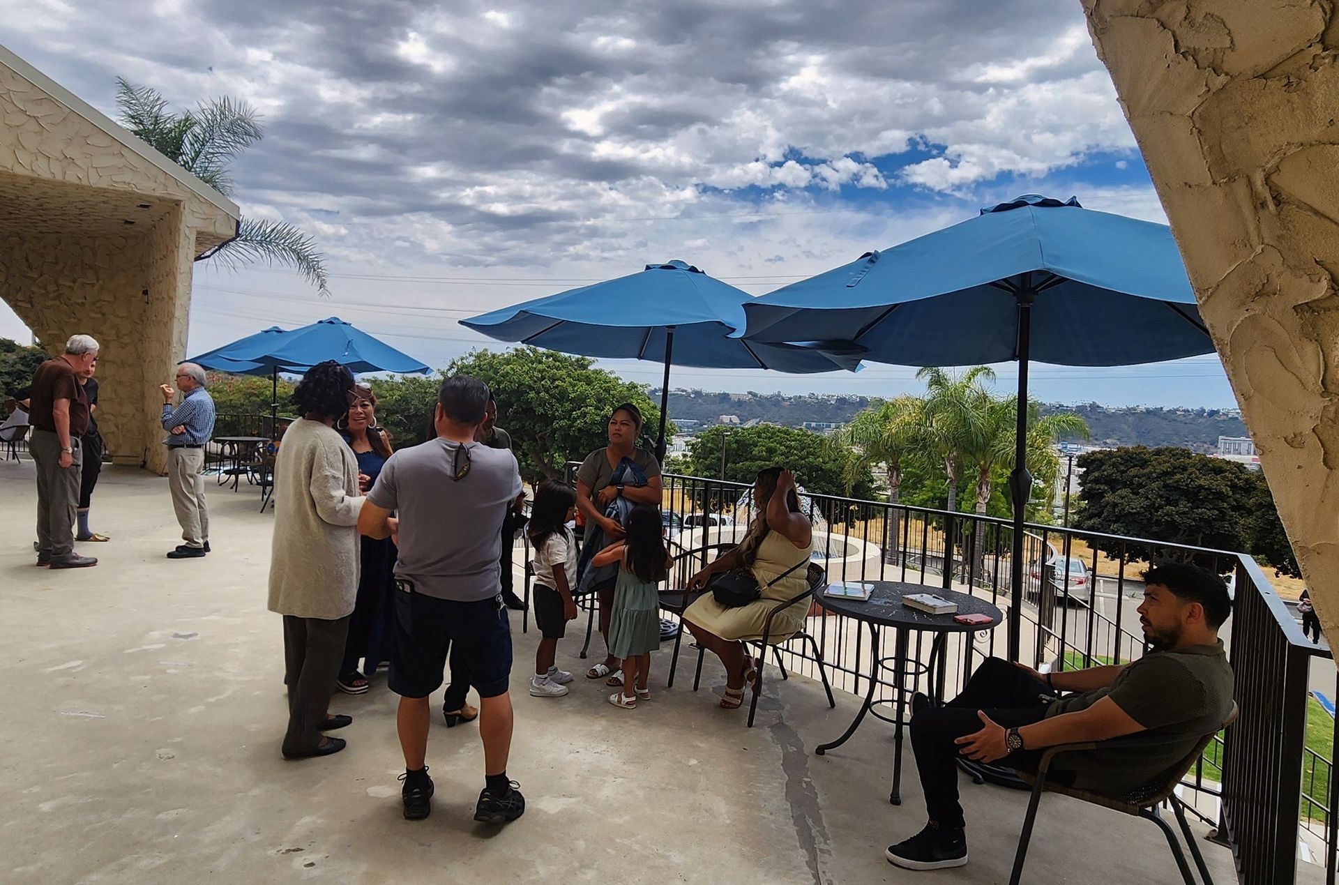 Group of people on a patio with blue umbrellas, overlooking a city on a cloudy day.