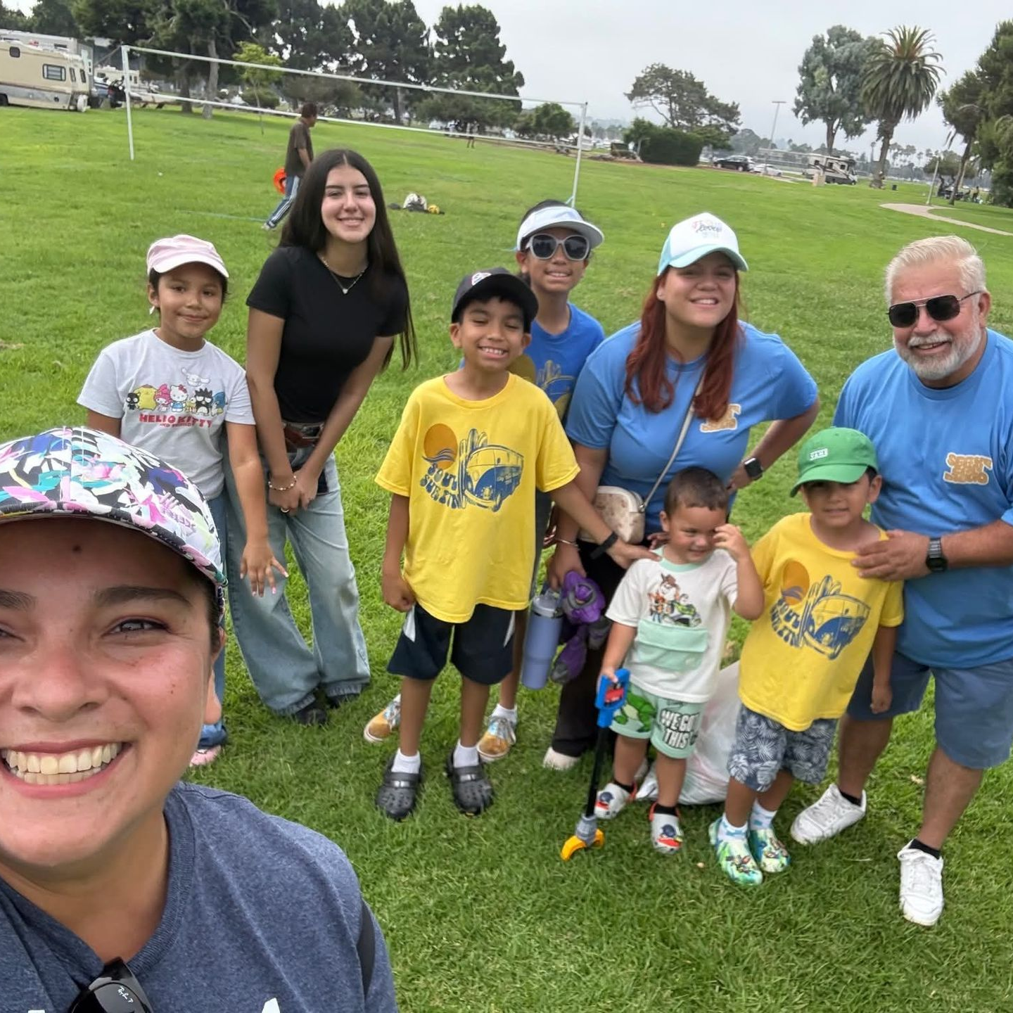 Group of people smiling on a grassy field. Several are wearing matching shirts. Sunny, outdoor setting.