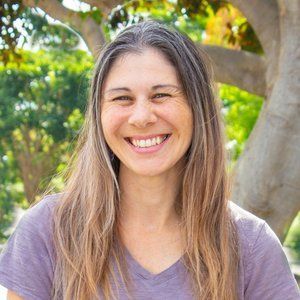 Woman with long gray-streaked hair smiles broadly, wearing a purple v-neck shirt outdoors in front of trees.