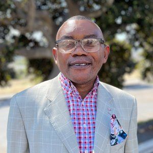 Man in a light-colored suit, red plaid shirt, glasses, and floral pocket square, smiling outdoors.