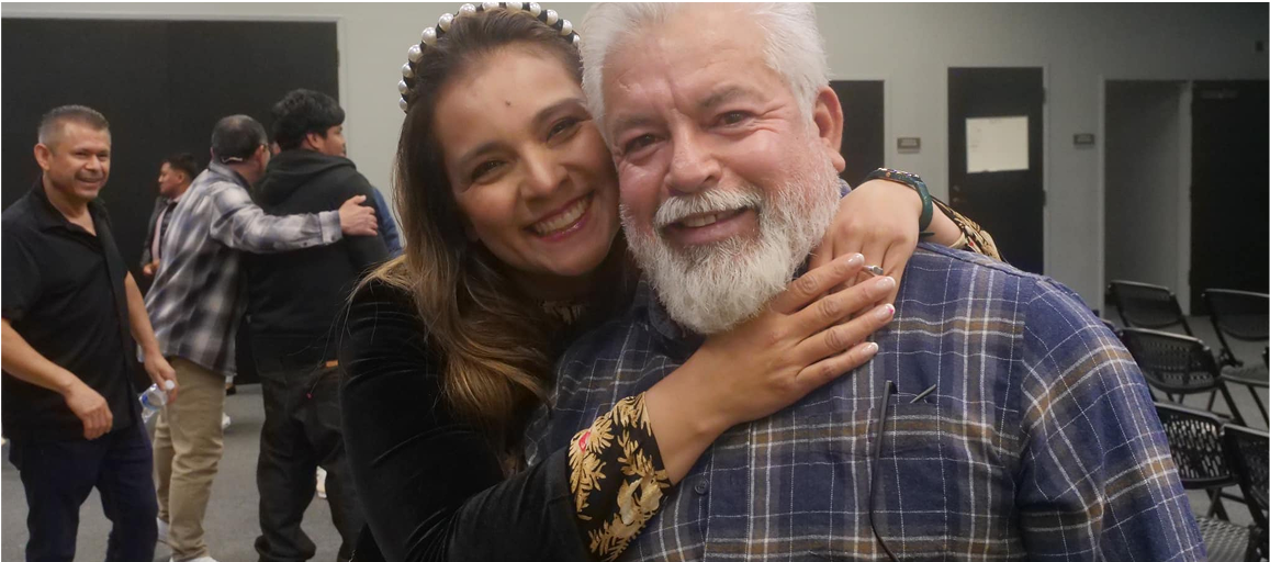 Woman hugging a smiling older man; others in the background. They are inside a building.