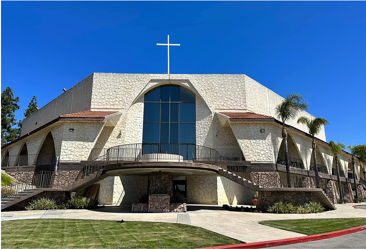 Church building with white cross on the roof and large arched window. Beige stone facade, blue sky, and green lawn.