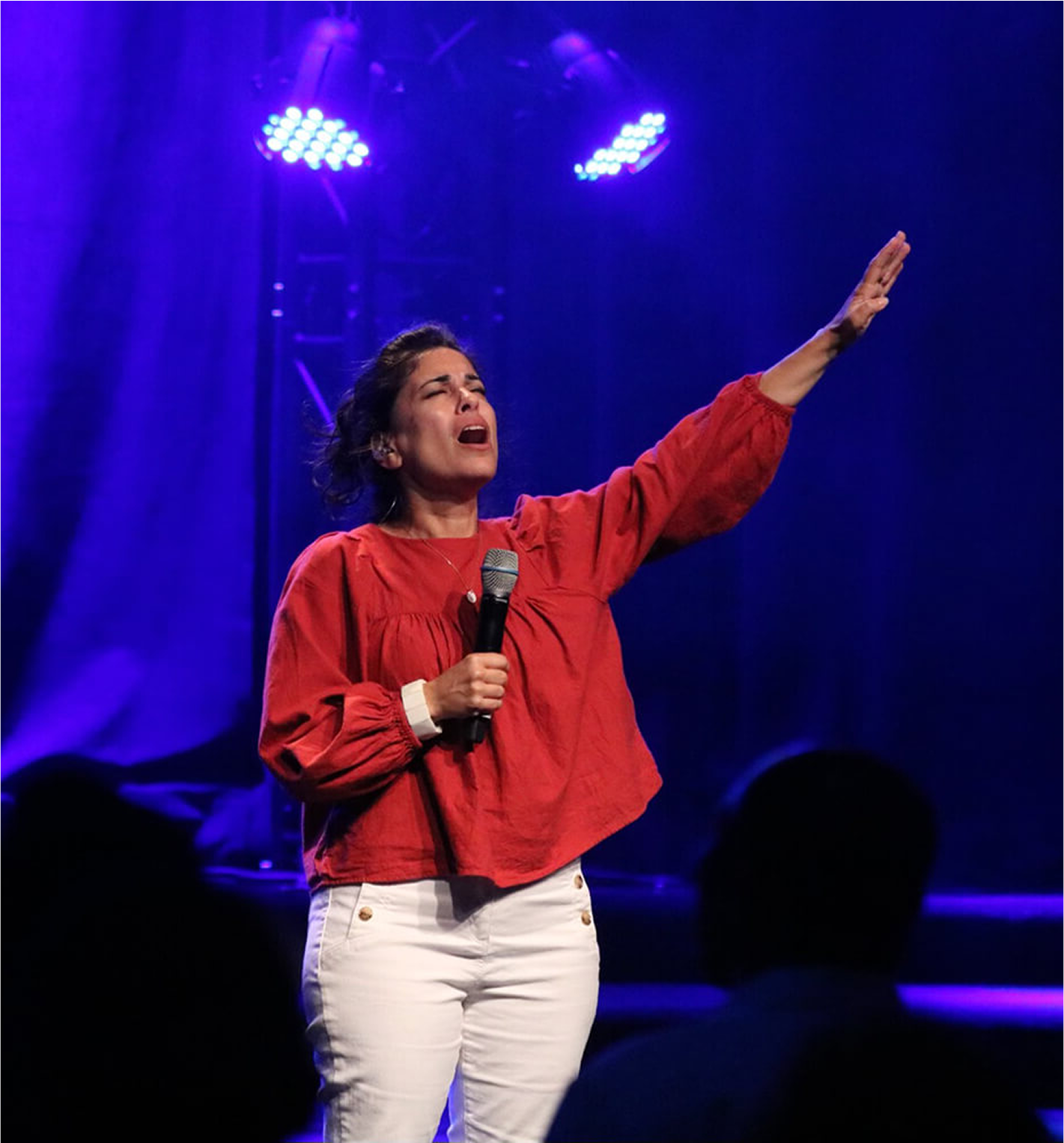 Woman on stage with microphone, arm raised, wearing a red shirt and white pants, under blue stage lights.