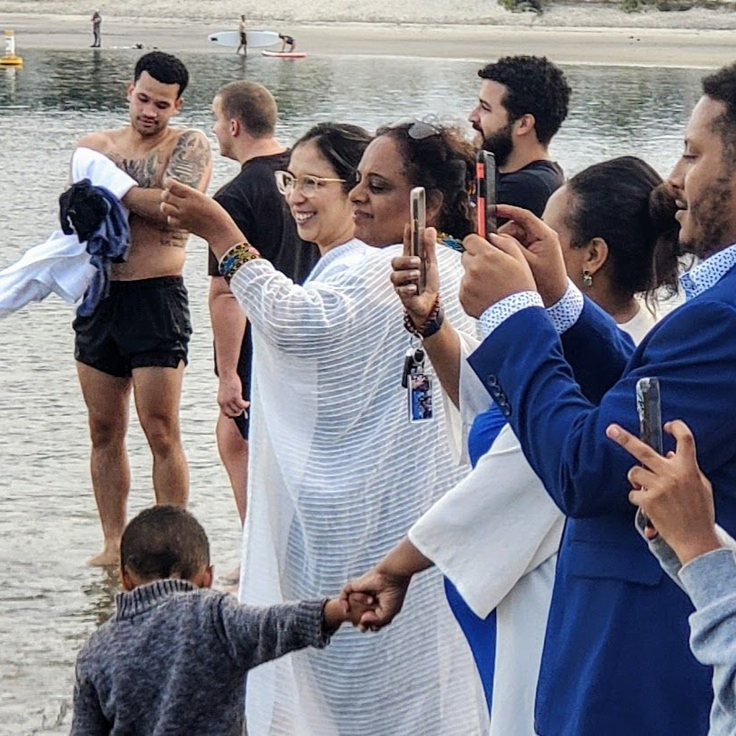 People taking photos on a beach; a man in water with tattoos; boy holds hands with a woman.