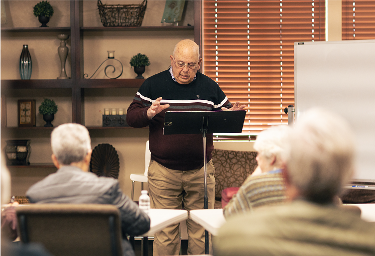 Man teaching a Bible class, gesturing, to audience in a room with bookshelves and a whiteboard.