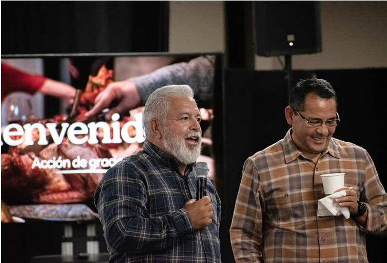 Two men in plaid shirts speaking in front of a screen with text. One holds a microphone, the other a cup. Hispanics. City View Espanol.