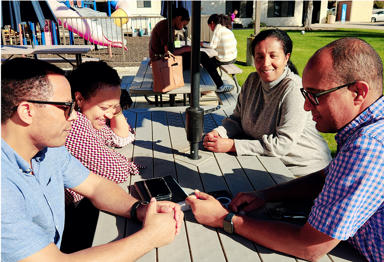 Four people sit around an outdoor table, talking. Two wear glasses. Sunny day, white building background.