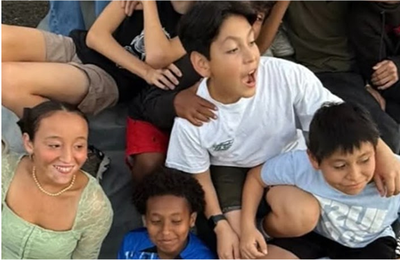 Group of kids smiling, sitting and leaning close together, one boy looks surprised.
