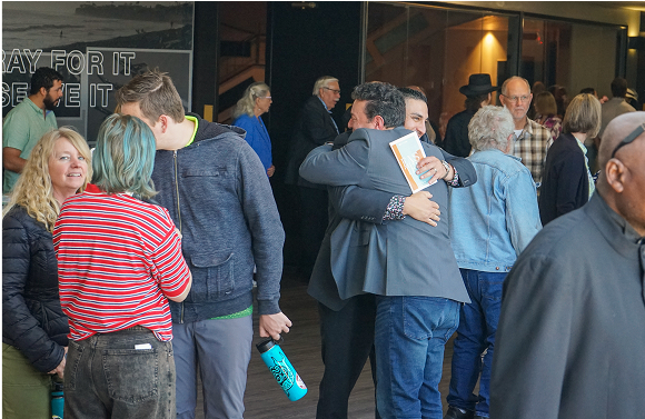 People embrace in a church. Several others watch. One holds a book.