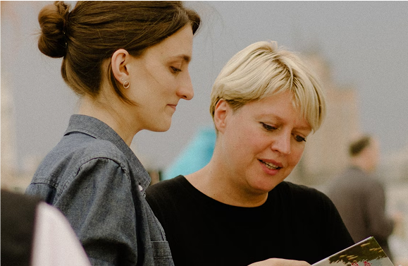 Two women looking at a brochure. The woman on the right has short blonde hair and is speaking.