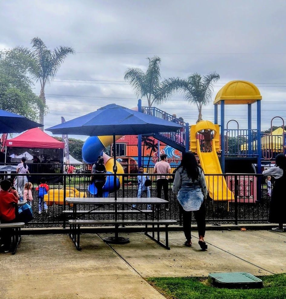 Playground scene with picnic tables, umbrellas, and people. A colorful play structure is in the background under a cloudy sky.