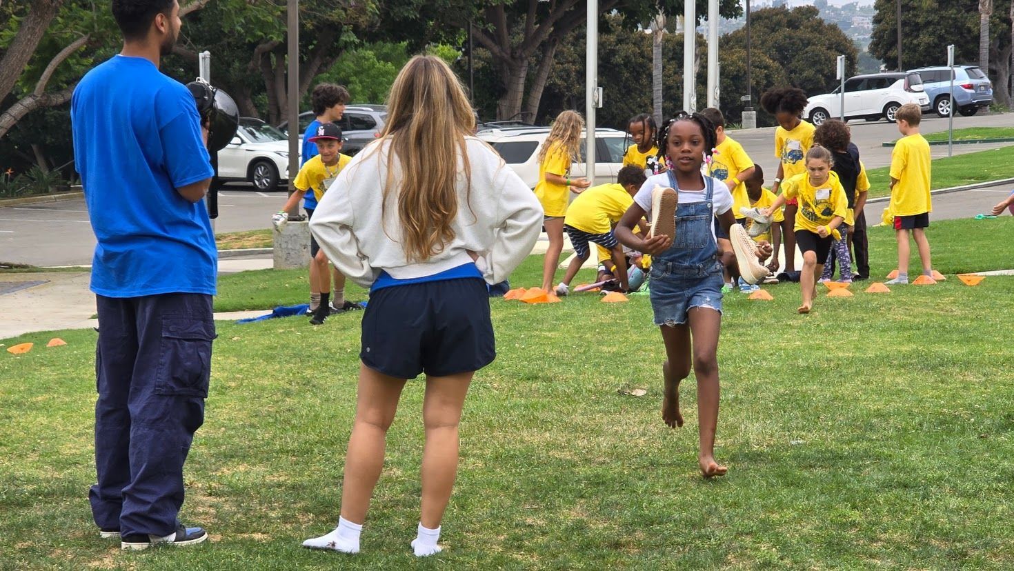 Girl running in a field; other children and adults watching, wearing yellow shirts.