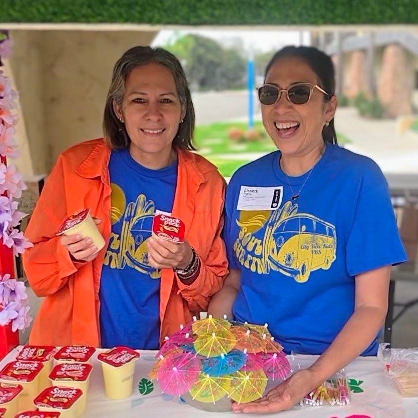 Two women smiling, holding snacks, wearing blue shirts, standing at a table decorated with cocktail umbrellas.
