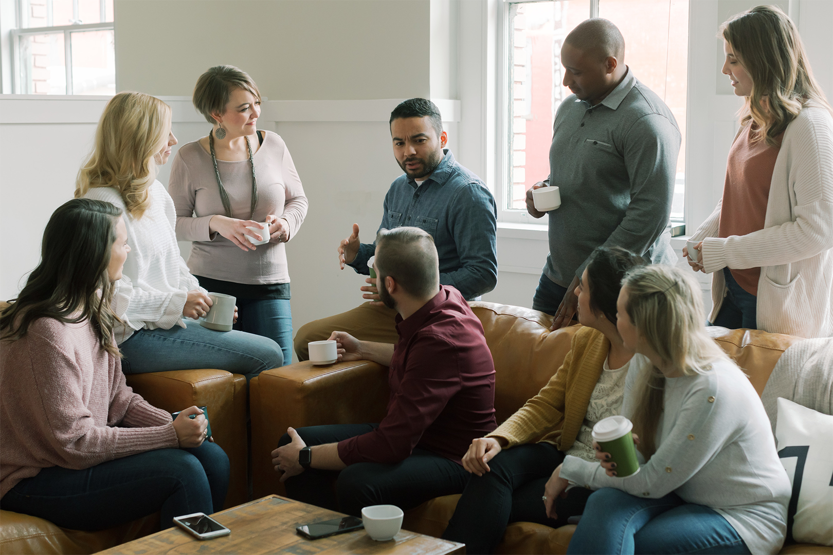 Group of people in a living room, some seated, some standing, conversing and holding mugs.