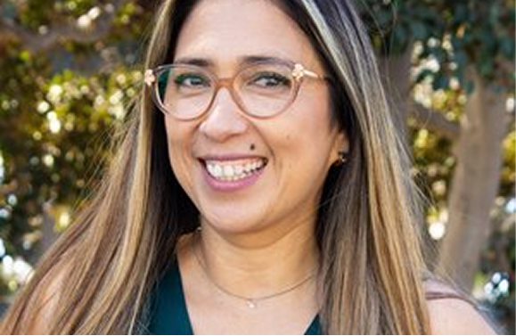 Woman with glasses smiles outdoors, with trees in the blurred background.