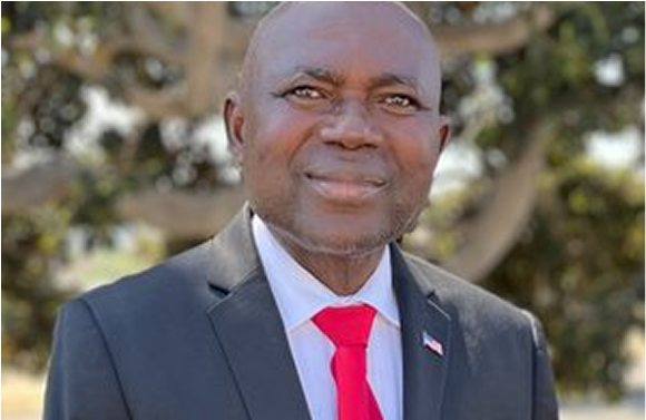 Man in black suit and red tie smiles, outdoors with a tree in the background.
