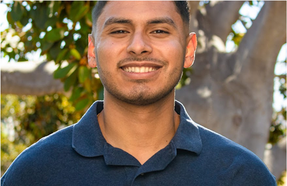 Man in blue polo shirt smiles outdoors, with trees in the background.