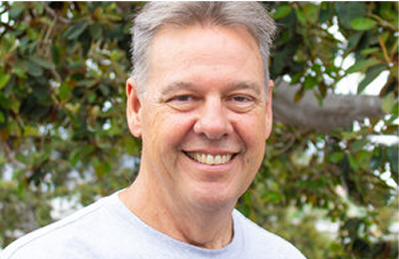Smiling man in light-colored shirt, outdoors in front of leafy green background.