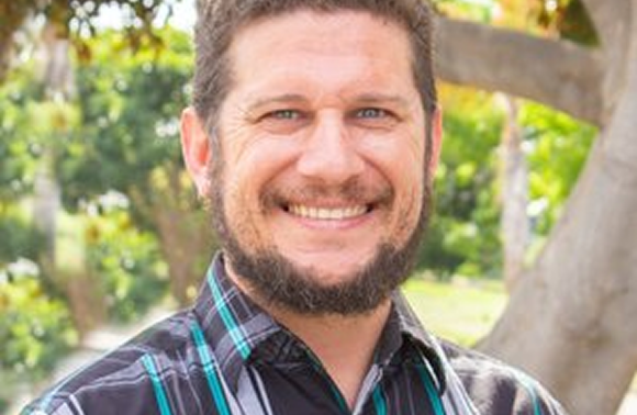Man with beard smiles at the camera, wearing a plaid shirt outdoors near trees.