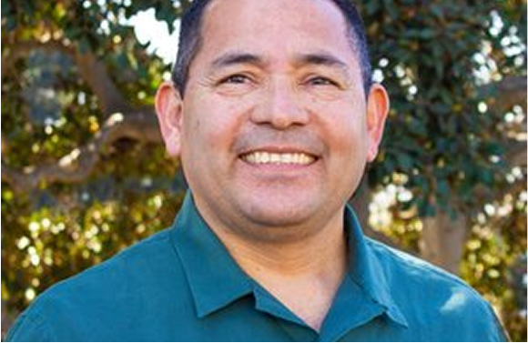 Man in green shirt smiles outdoors, blurred trees in background.