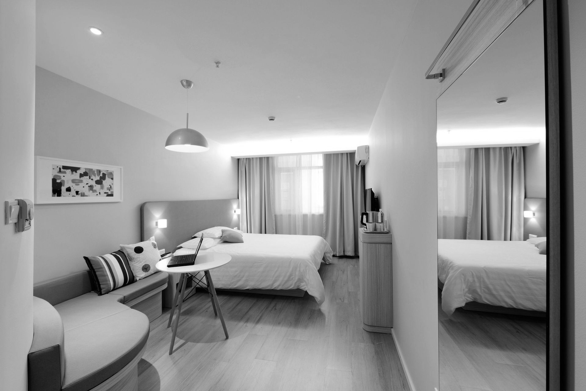 Monochrome hotel room with bed, sofa, table, mirror, and window.