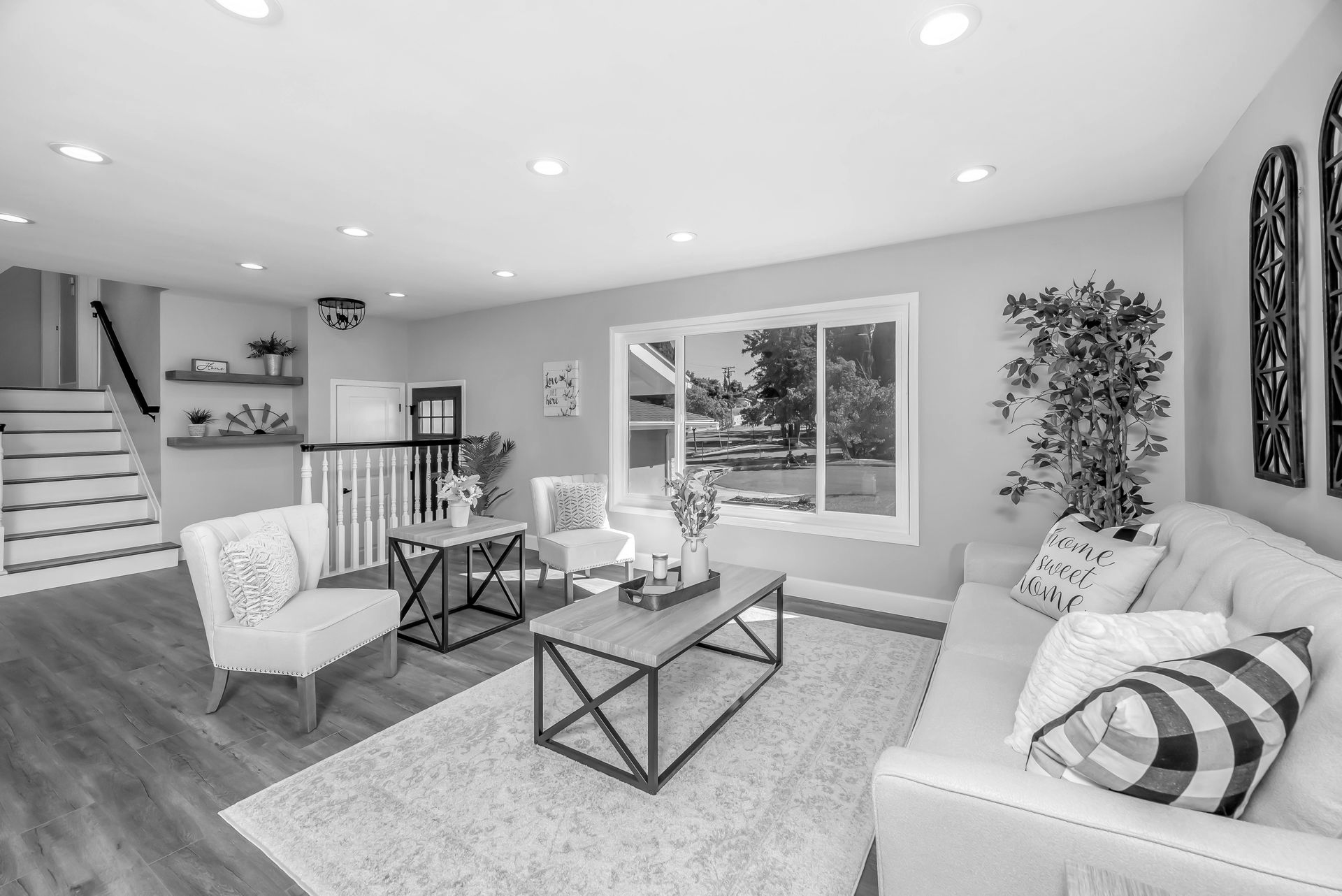 Living room with white sofa, chairs, coffee tables, window, and stairs in grayscale.