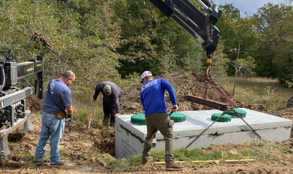 Three workers prepare to lower a concrete septic tank into an excavated pit in a wooded area using a crane.