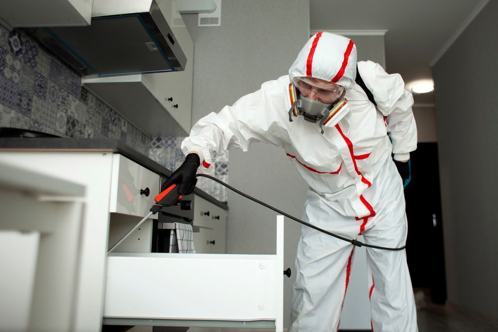 A man in a protective suit is spraying a drawer in a kitchen.