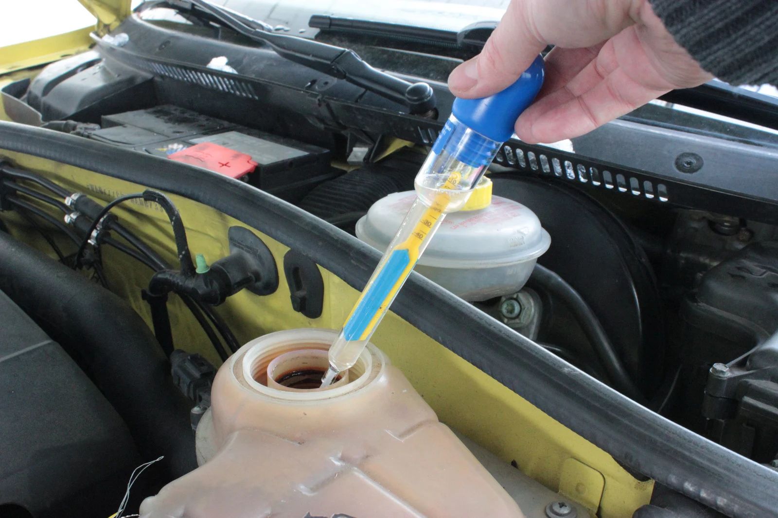 Person checking coolant level in a car's reservoir with a tester. Yellow and blue tool, engine bay.