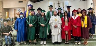A diverse group of graduates in caps and gowns pose with a priest. 