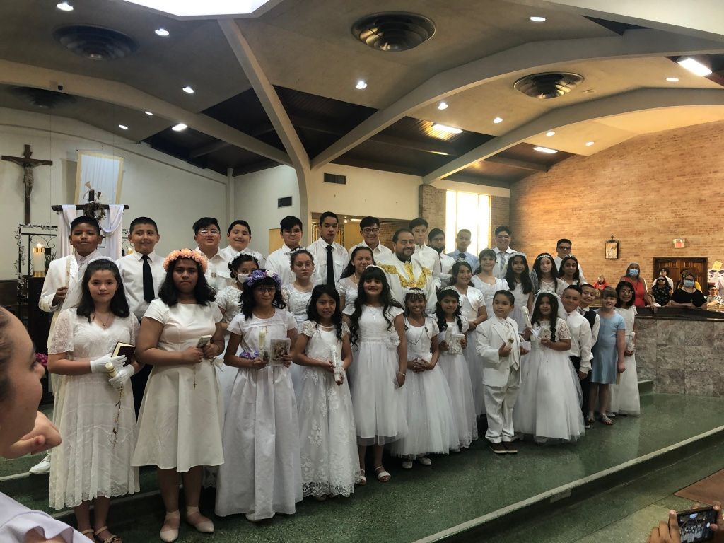 A group of children and teens in white dresses and suits stand together in a church interior, smiling. 