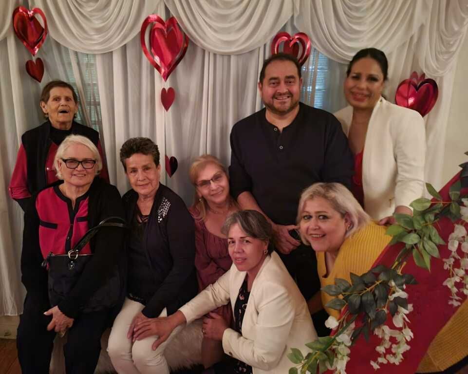 A group of people pose in front of a white curtain decorated with red heart balloons. 