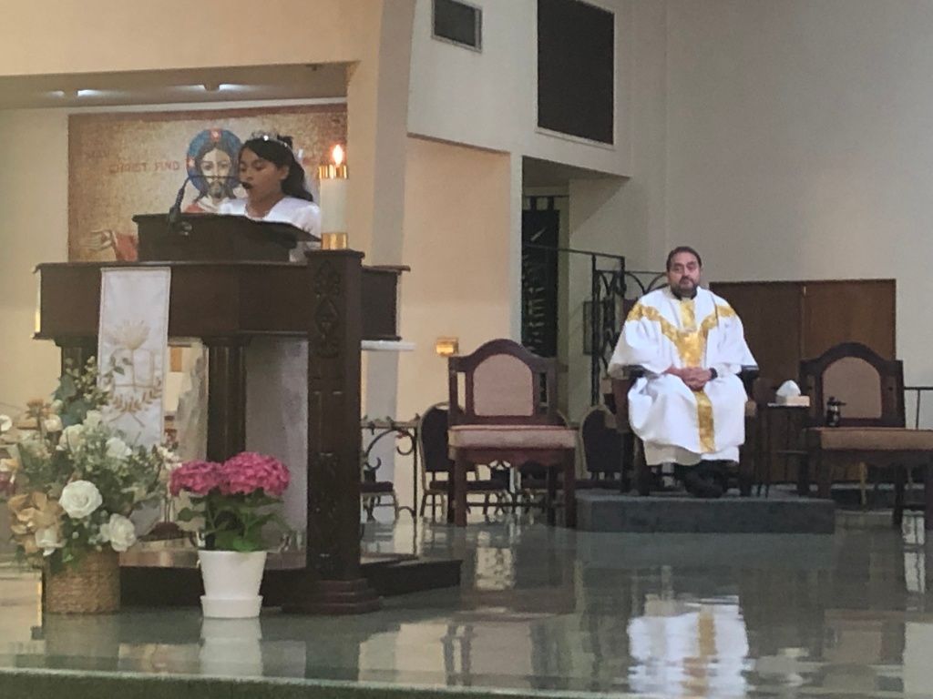 A young girl stands at a church lectern, reading and a priest in a white robe with gold accents sits behind her. 