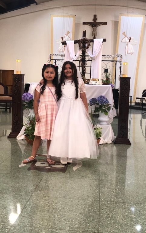 Two young girls smiling in a church, one in a white dress and the other in pink plaid.