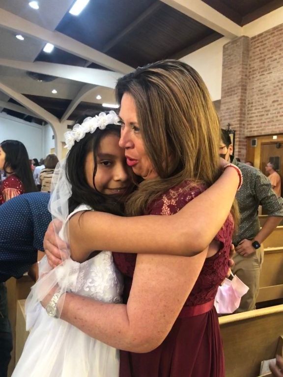 Girl in a white dress hugs a woman in a red gown inside a church.