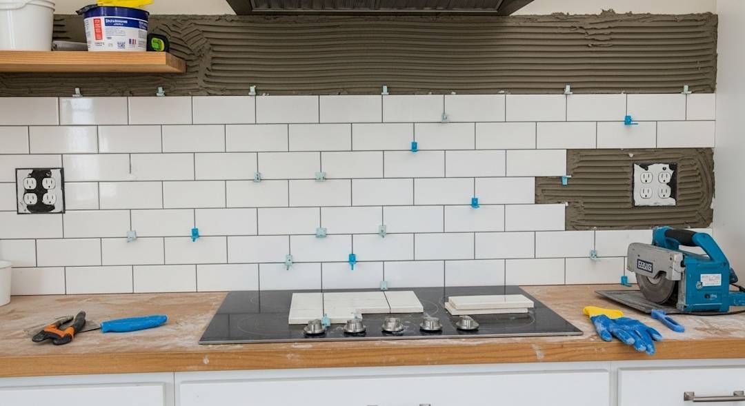 Kitchen backsplash tile being installed above a countertop, with tools, spacers, and a stove centered below.