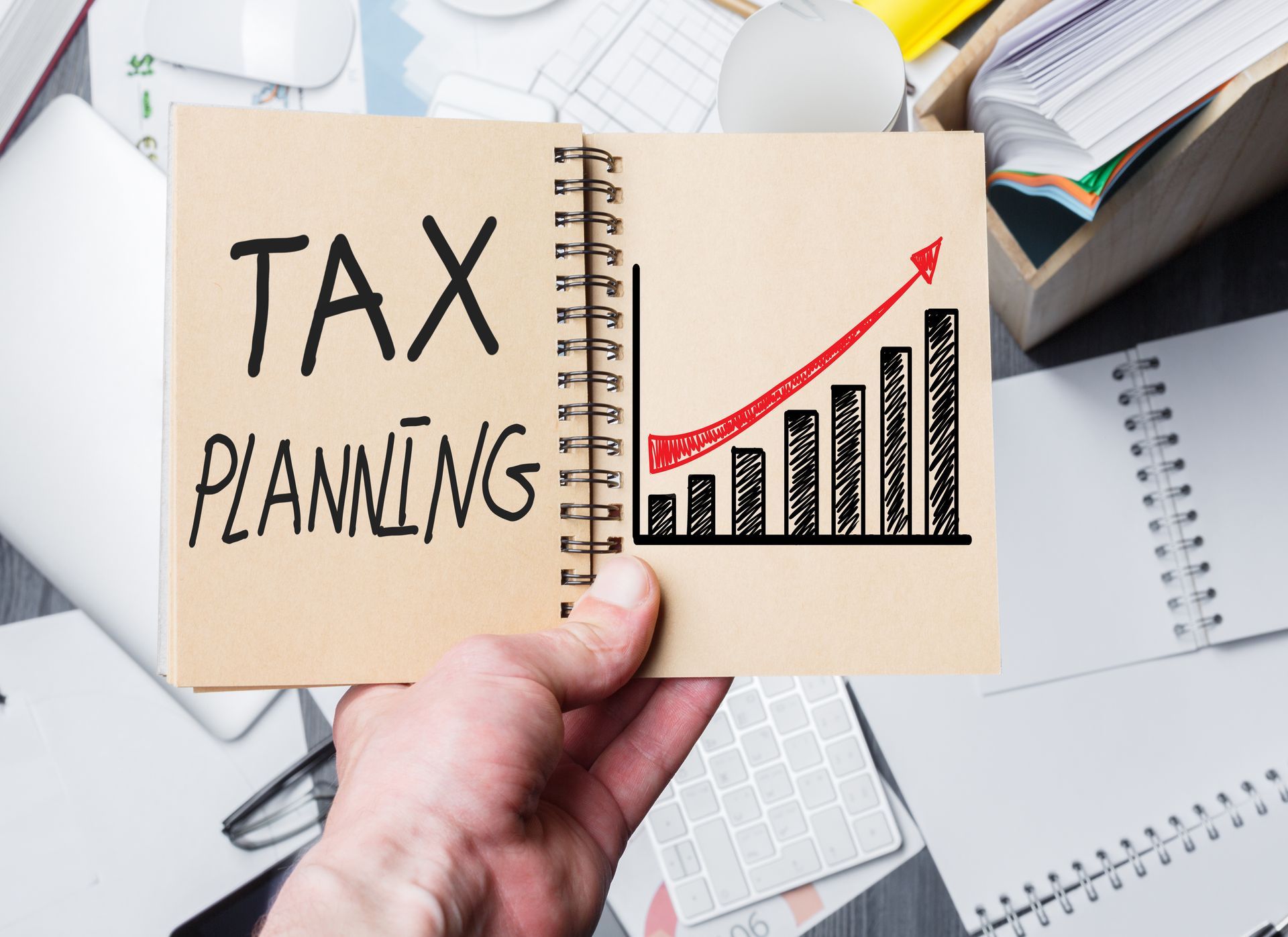 Hand holding notebook labeled “Tax Planning” with rising bar and arrow chart on desk Hand holding notebook labeled “Tax Planning” with rising bar and arrow chart on desk