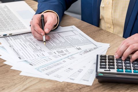Hands working on tax forms beside a calculator and laptop on a desk