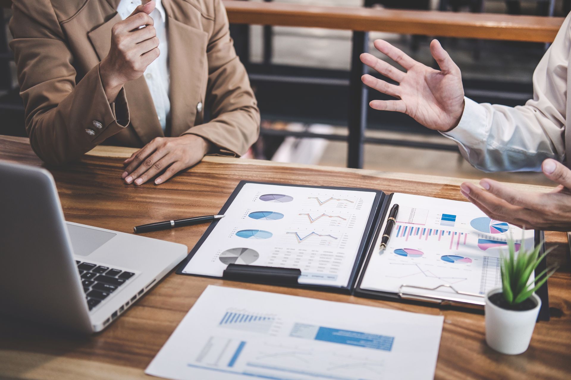 Business meeting with laptop, charts, and two people discussing reports at a desk Business meeting with laptop, charts, and two people discussing reports at a desk