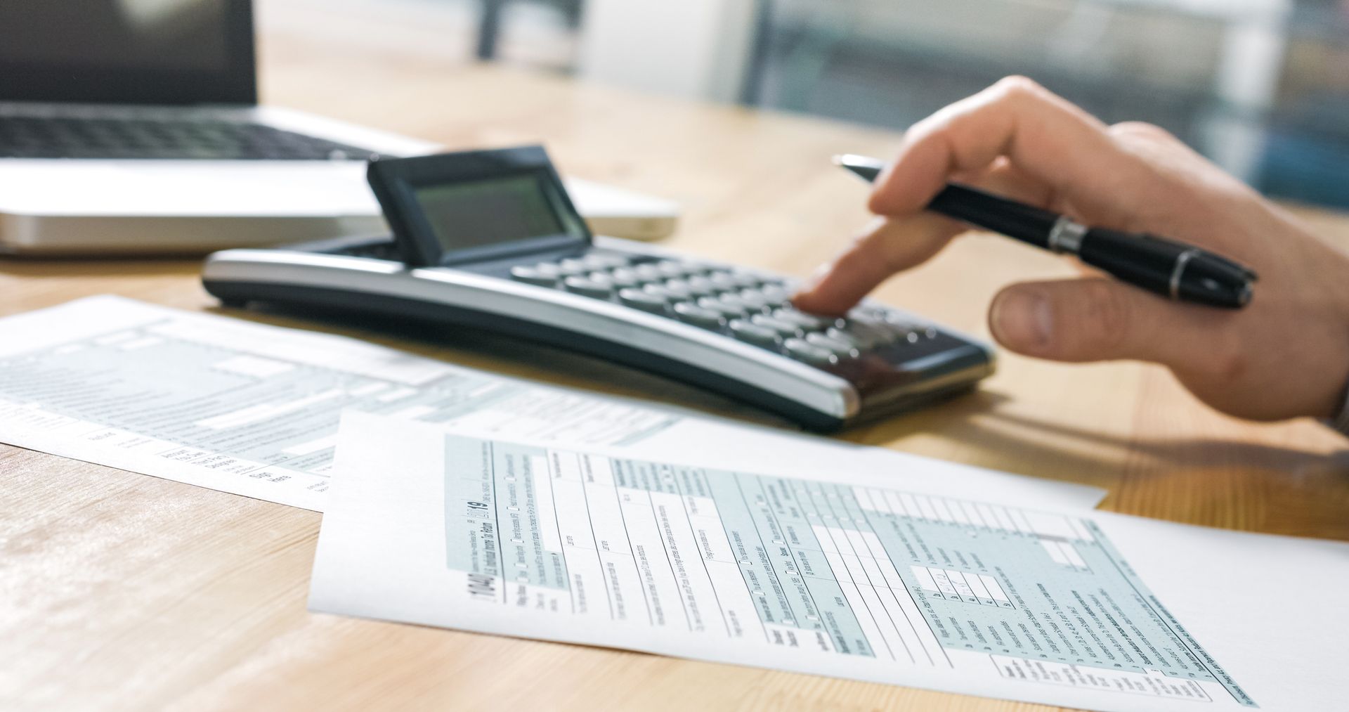 Hand using a calculator beside paperwork on a desk, with a laptop in the background Hand using a calculator beside paperwork on a desk, with a laptop in the background