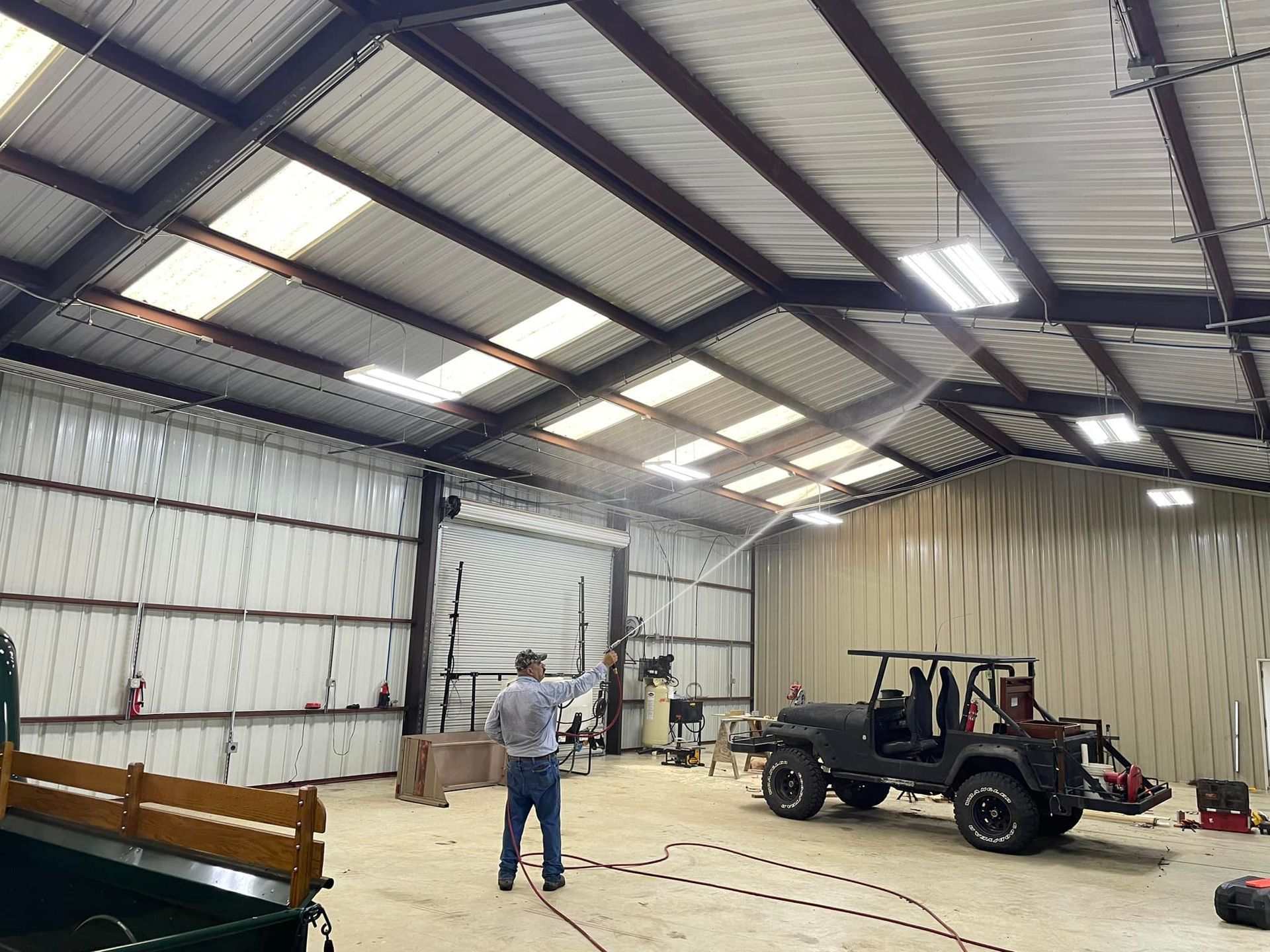 Spacious metal workshop with a person standing near a black Jeep and a red trailer under bright roof lights