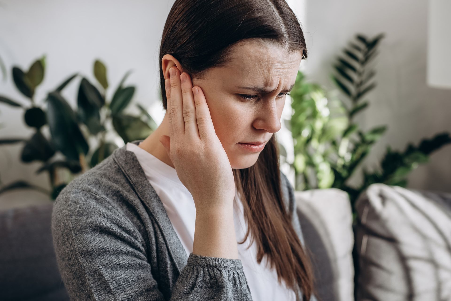 A person with a pained expression holding their hand against their ear in a living room with plants.