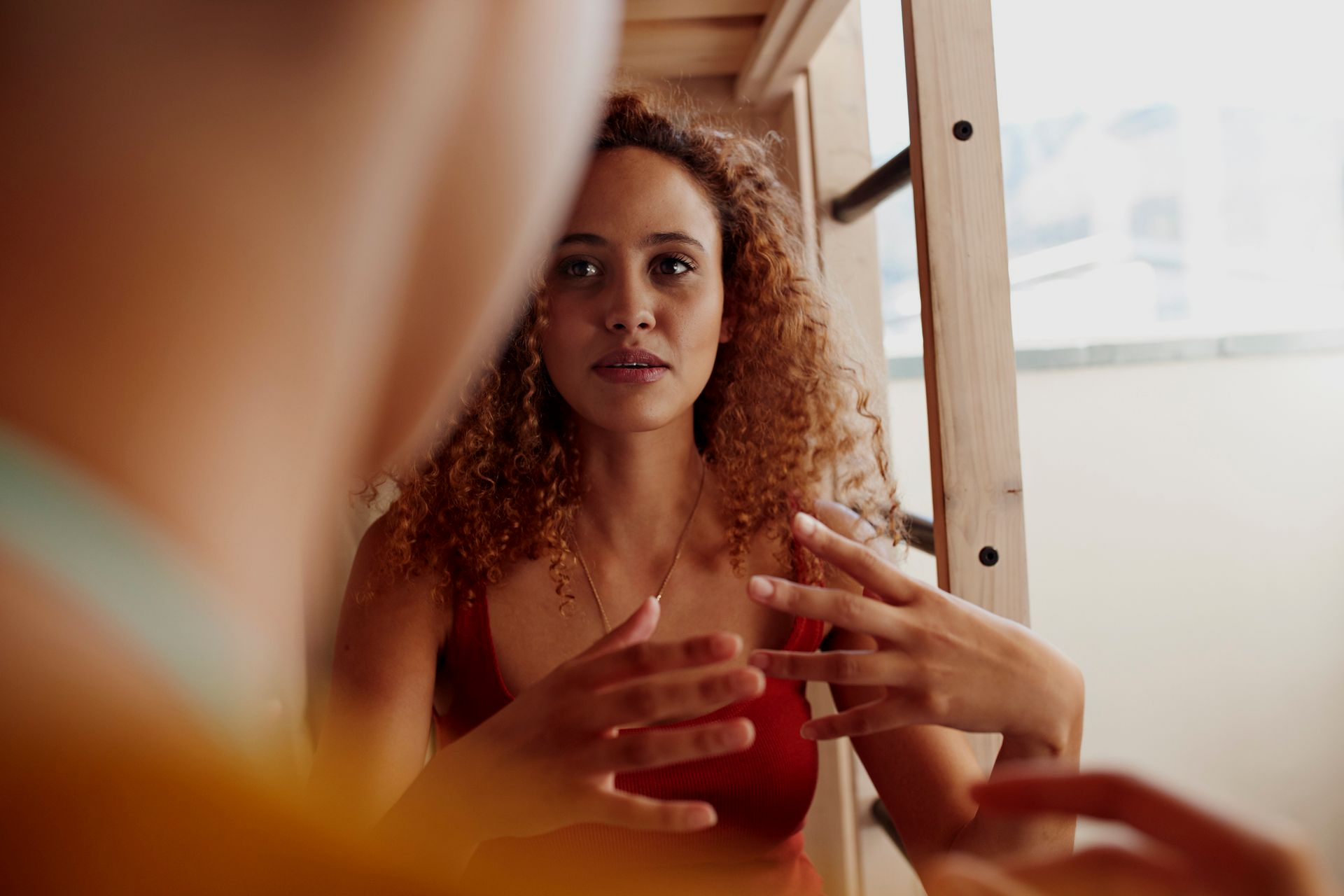 A person with curly hair wearing a red top gestures while talking to someone off-camera in a casual setting.