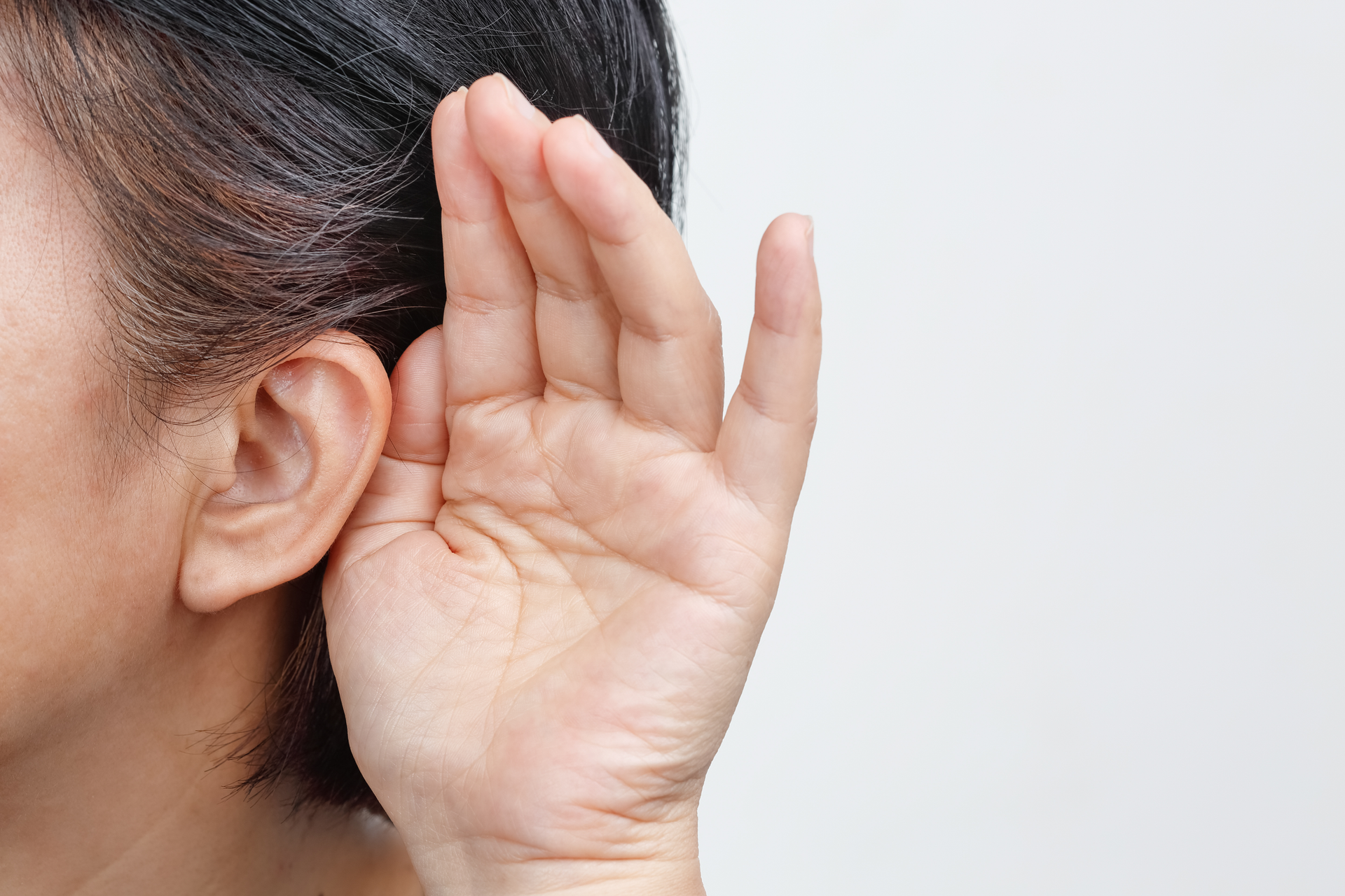 A person cupping their hand behind their ear to hear better against a plain light background.