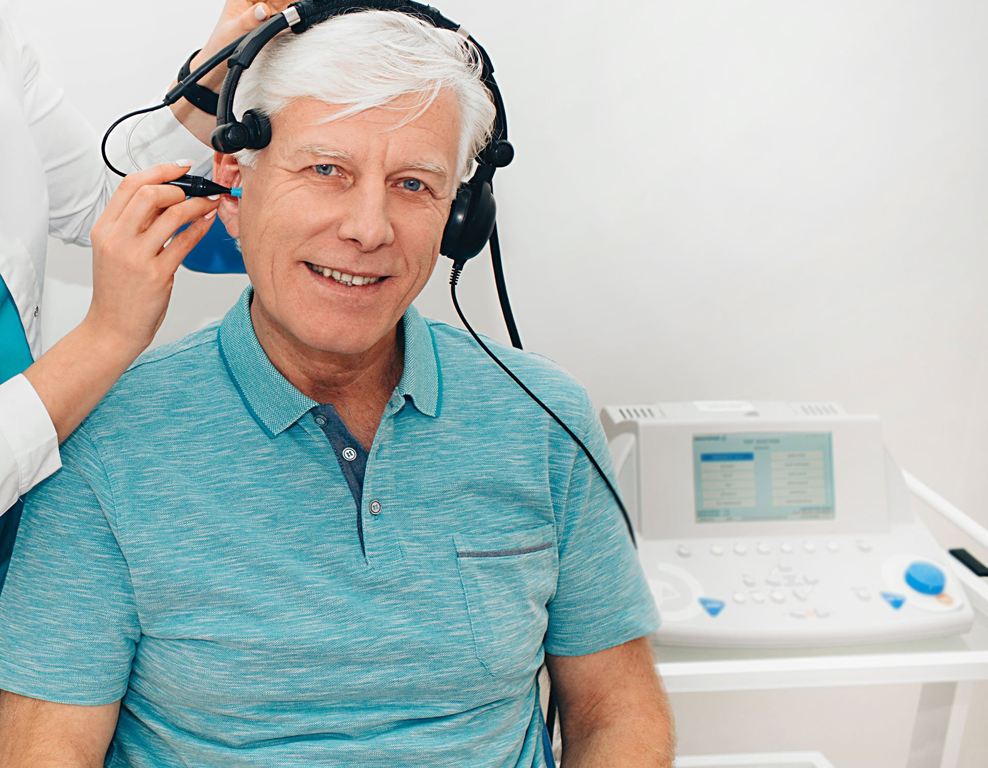 Man in turquoise shirt gets a hearing test. Doctor adjusts headphones in a clinic.