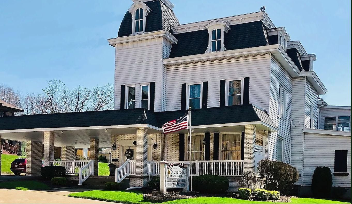 Large white Victorian-style house with striped awnings, front porch, and manicured lawn on a sunny day